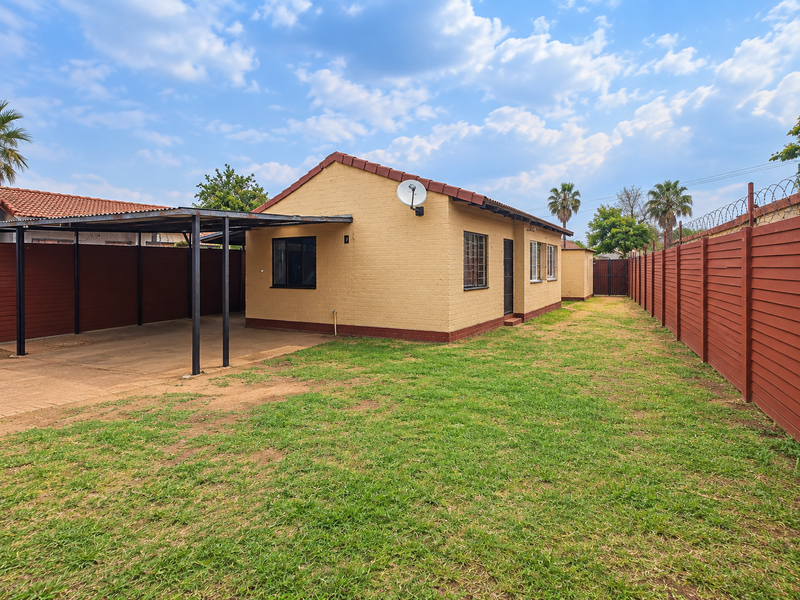 Rear view of the property featuring a carport, brick boundary walls with security fencing, and a spacious garden area.