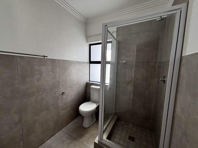 A clean, modern bathroom featuring a glass-enclosed shower, neutral wall and floor tiling, and a standard toilet.