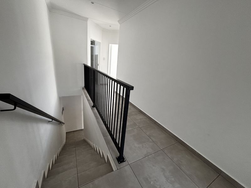 A clean, modern hallway featuring tiled flooring, a black metal balustrade, and a staircase leading to the lower level.