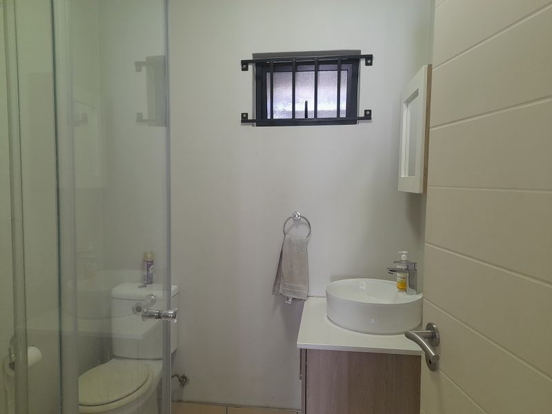 A compact, modern bathroom featuring a glass-enclosed shower, a vanity with a vessel sink, and a window with security bars.