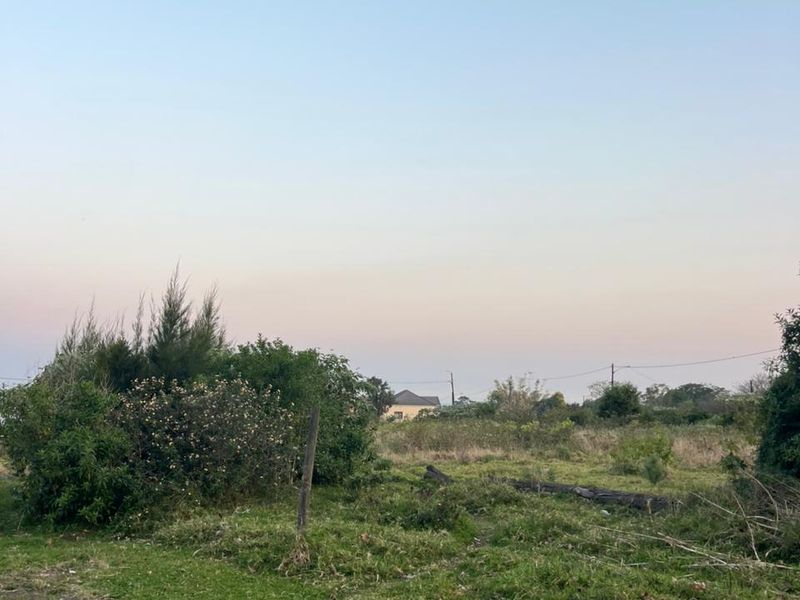 A view of a vacant residential plot featuring natural grass, scattered shrubbery, and a perimeter fence post.