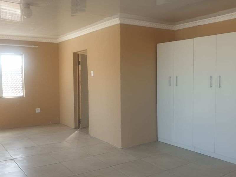 A spacious, empty bedroom featuring neutral wall tones, tiled flooring, a glossy ceiling with decorative cornice, and a large white built-in wardrobe.