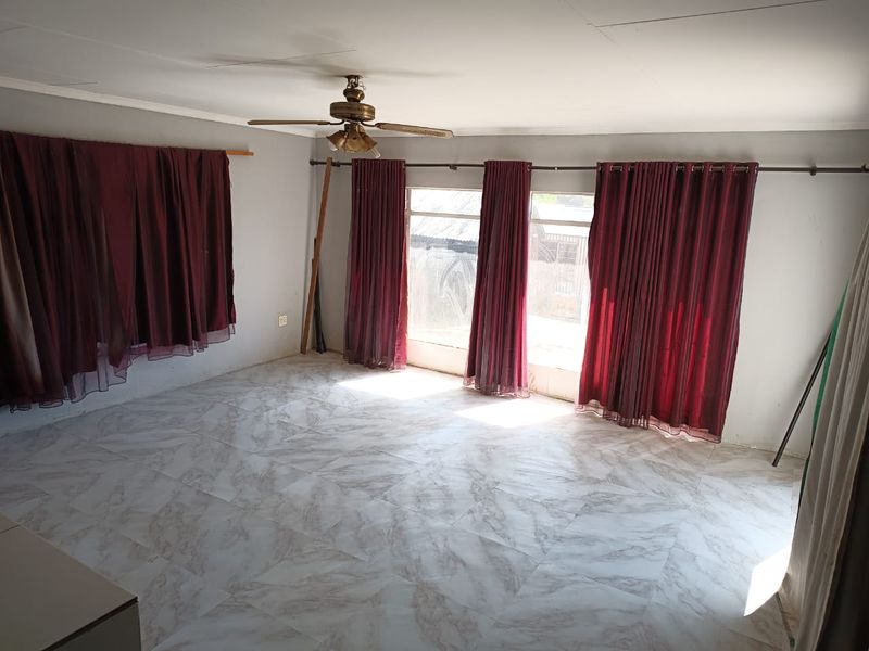 A spacious, empty living room featuring tiled flooring, a ceiling fan, and large windows with installed curtain rods.