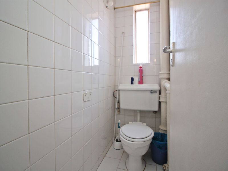 A compact bathroom featuring white ceramic wall and floor tiles, a standard toilet, and an exposed plumbing pipe.