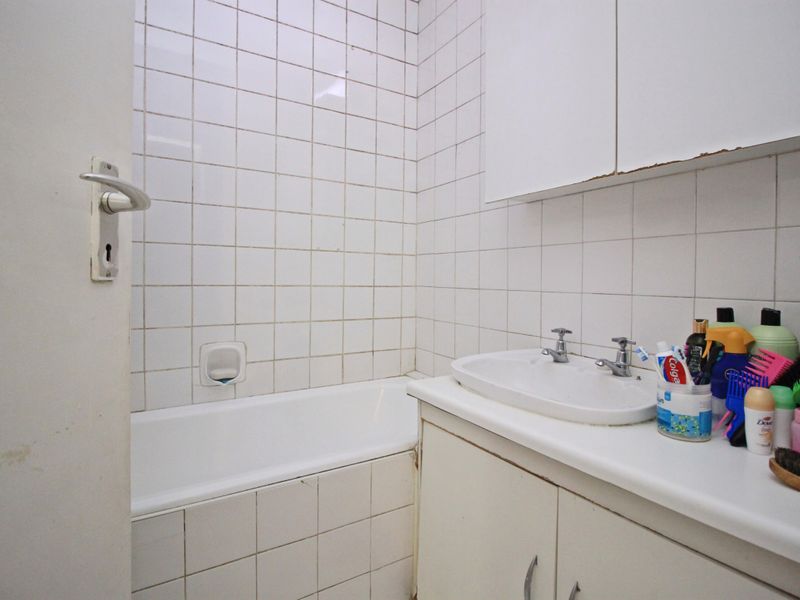 A compact bathroom featuring a built-in bathtub, vanity cabinet with washbasin, and wall-mounted storage, finished with white ceramic wall tiles.