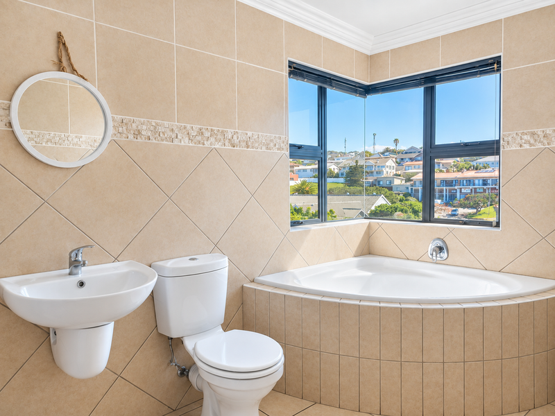 A bright, well-maintained bathroom featuring a corner bathtub, wall-mounted basin, and dual-flush toilet, complemented by neutral ceramic tiling and a scenic corner window.