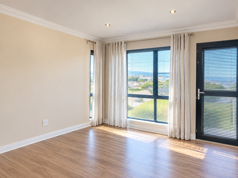 A bright, empty bedroom featuring modern laminate flooring, recessed downlights, and large windows with venetian blinds and access to an outdoor area.