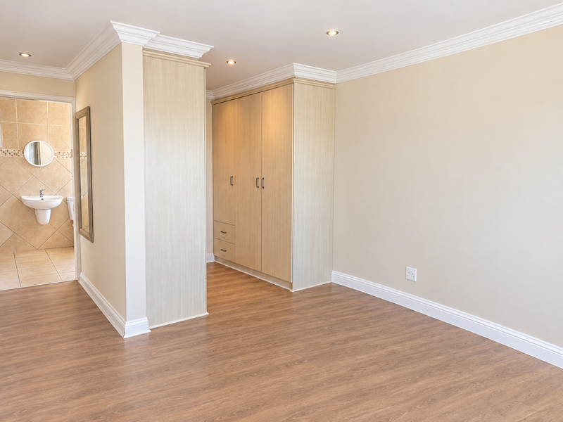 A bright, empty bedroom featuring light wood-finish built-in cupboards, laminate flooring, crown molding, and an adjoining en-suite bathroom.