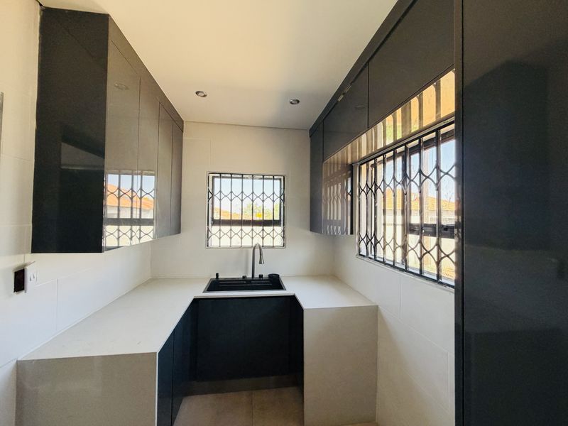 A modern, minimalist scullery featuring sleek dark cabinetry, white stone countertops, an undermount sink, and security bars on the windows.