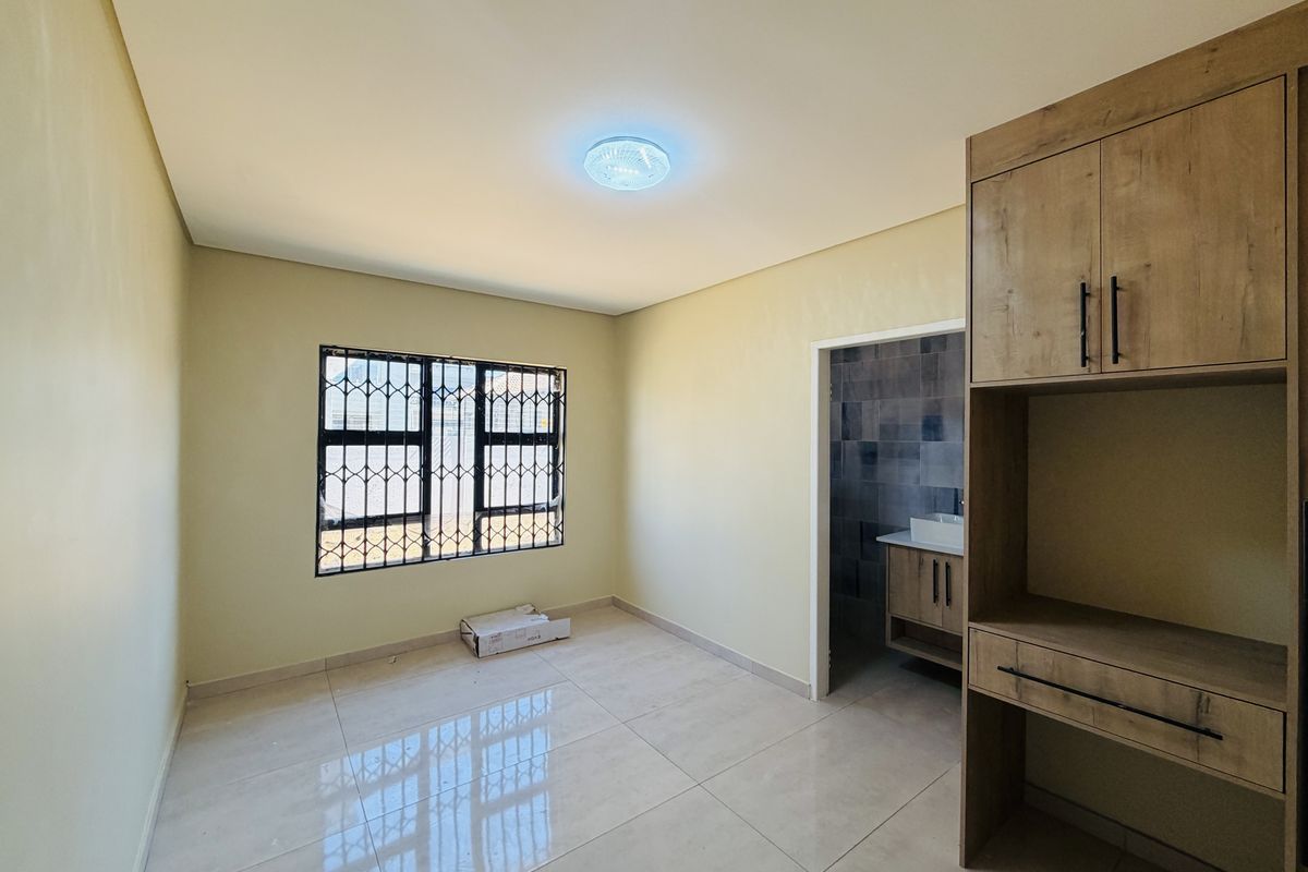 A bright, empty bedroom featuring tiled flooring, built-in cabinetry, and a window with security bars, leading into an en-suite bathroom area.