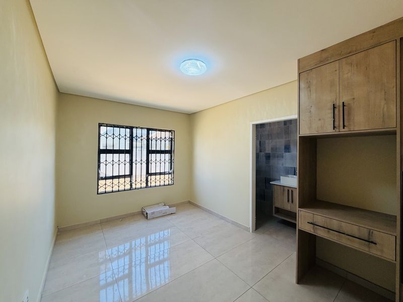 A bright, empty bedroom featuring tiled flooring, built-in cabinetry, and a window with security bars, leading into an en-suite bathroom area.