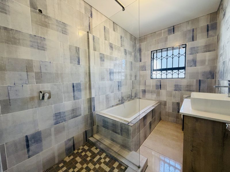 Modern bathroom featuring a built-in bathtub, glass shower screen, and vanity basin with decorative tiled walls and security bars on the window.