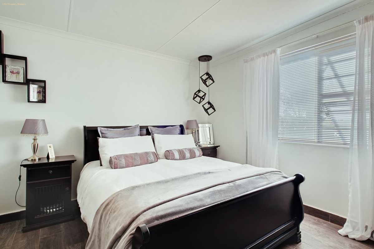 A well-maintained bedroom featuring dark laminate flooring, neutral walls, and a modern pendant light fixture with natural light provided by a window with blinds.