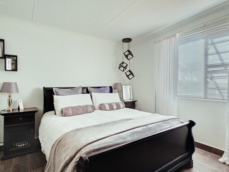 A well-maintained bedroom featuring dark laminate flooring, neutral walls, and a modern pendant light fixture with natural light provided by a window with blinds.