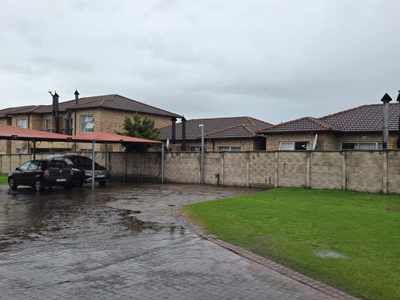 Exterior view of the apartment complex featuring a paved driveway, covered carport parking, and a brick boundary wall with a landscaped lawn area.