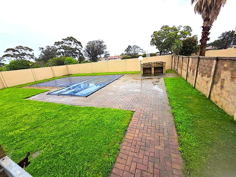 Outdoor pool area featuring a swimming pool with cover, brick paved patio, built-in braai, and surrounding lawn enclosed by a perimeter wall.