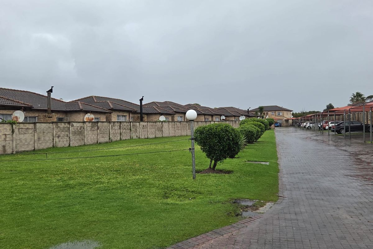 Exterior view of the apartment complex featuring a paved driveway, covered parking bays, and a shared lawn area with a boundary wall.