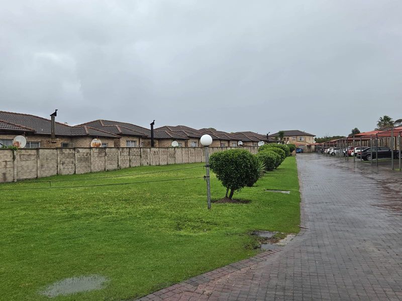 Exterior view of the apartment complex featuring a paved driveway, covered parking bays, and a shared lawn area with a boundary wall.