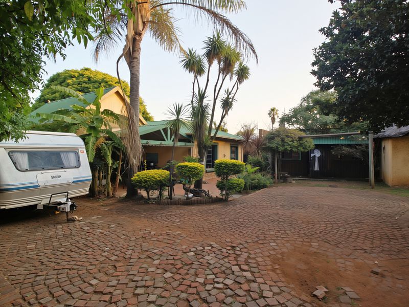 A spacious rear exterior area featuring a large brick-paved driveway, mature landscaping, and a covered patio attached to the main dwelling.