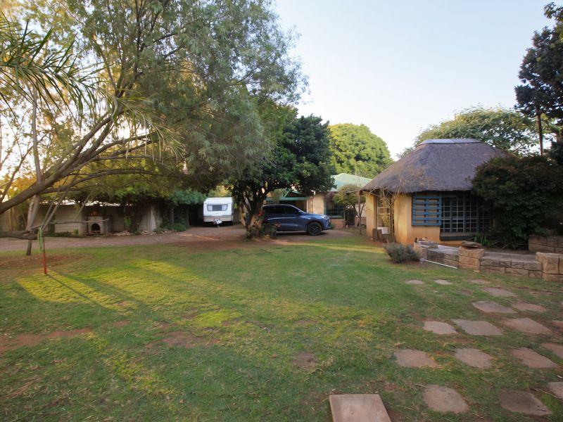 A spacious garden area featuring a thatched-roof structure, stone retaining wall, and paved pathways, set within a mature, tree-lined residential property.