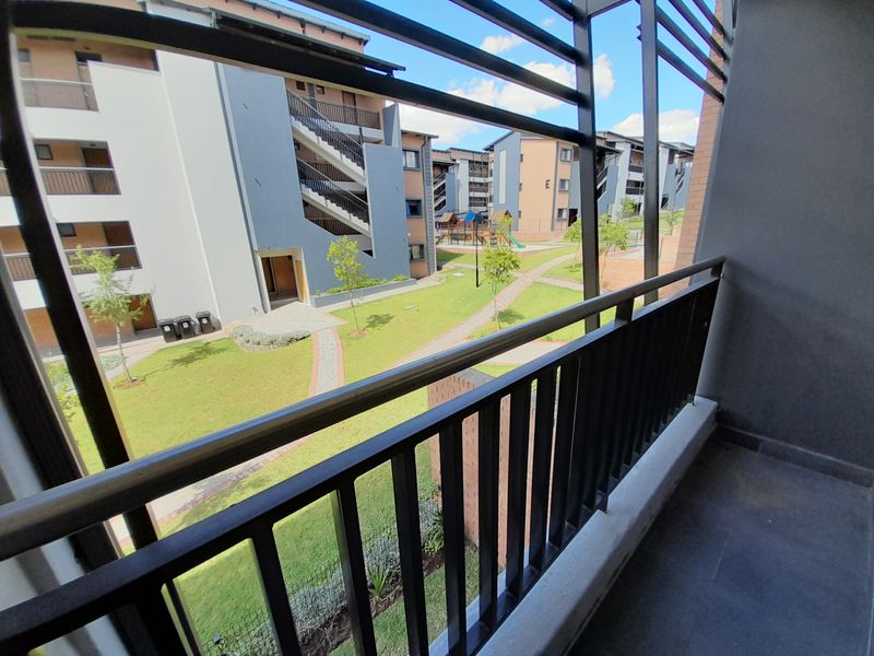 A private balcony overlooking a well-maintained communal garden area with paved pathways and modern apartment building architecture.
