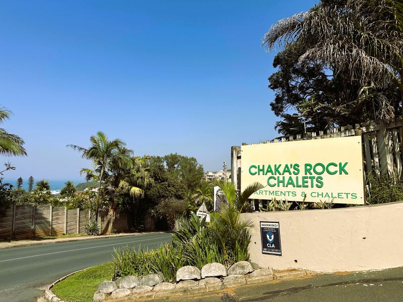 Street view of the Chaka's Rock Chalets entrance, featuring signage, boundary wall, and established coastal landscaping.
