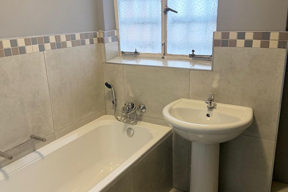 A clean, functional bathroom featuring a pedestal basin, a built-in bathtub with a handheld shower mixer, and neutral wall tiling.