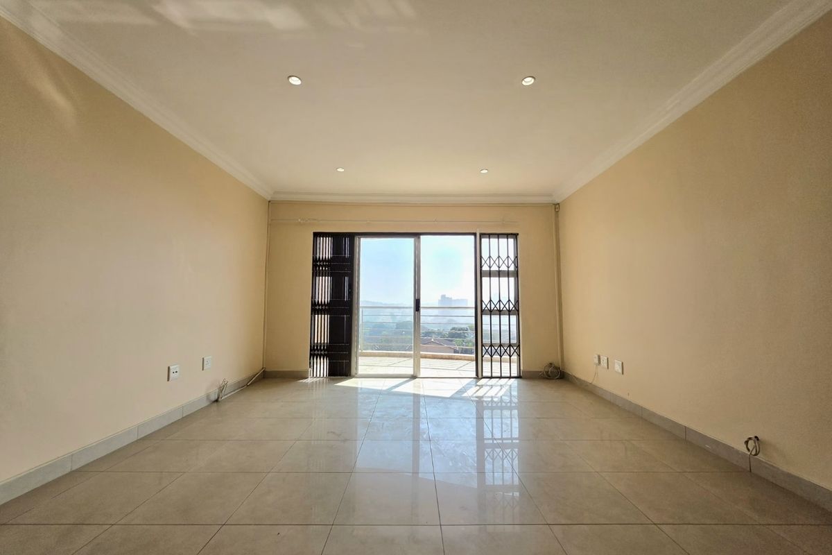 A bright, empty living room featuring neutral tiled flooring, recessed downlights, and a sliding glass door leading to an outdoor balcony.