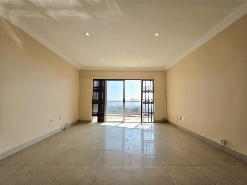 A bright, empty living room featuring neutral tiled flooring, recessed downlights, and a sliding glass door leading to an outdoor balcony.