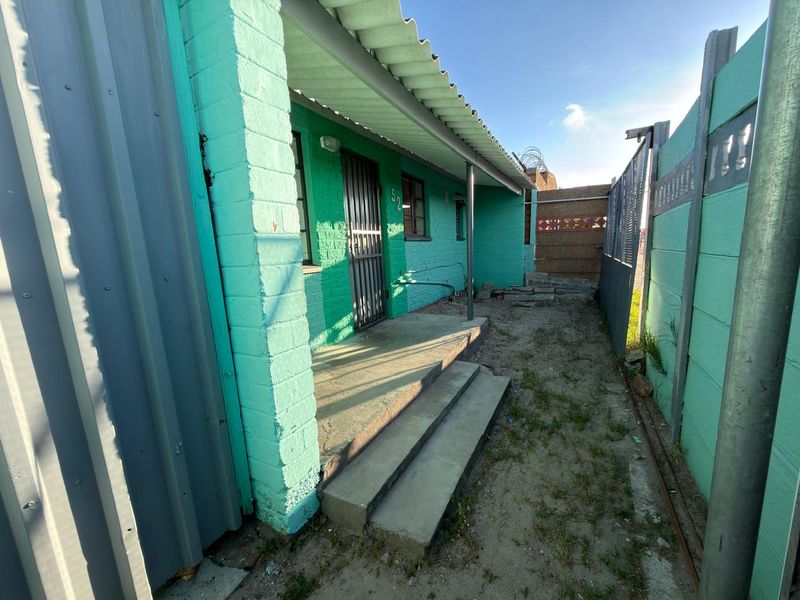 A side view of the property showing a concrete entrance path, security gate, and perimeter walling with a corrugated metal roof overhang.