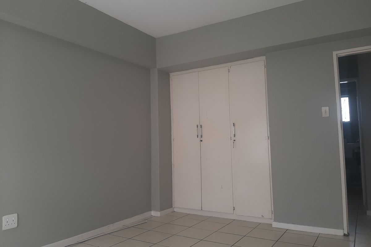 A clean, empty bedroom featuring neutral grey walls, tiled flooring, and a built-in white wardrobe with access to an adjacent room.