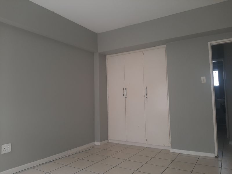 A clean, empty bedroom featuring neutral grey walls, tiled flooring, and a built-in white wardrobe with access to an adjacent room.
