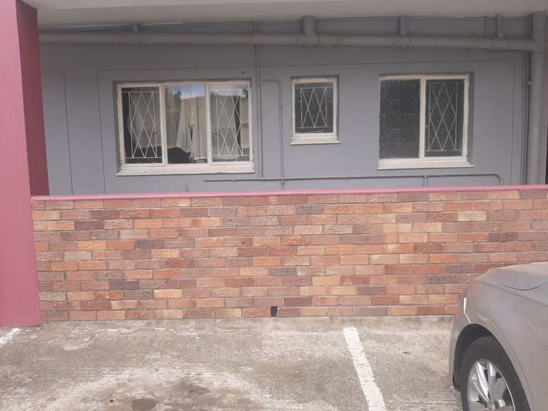 Exterior view of a townhouse facade featuring a brick boundary wall, security-barred windows, and exposed exterior plumbing pipes above a concrete driveway.