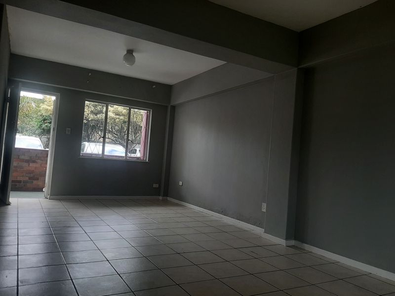 An empty, open-plan living area featuring neutral tiled flooring, grey painted walls, and natural light from a window with security bars.