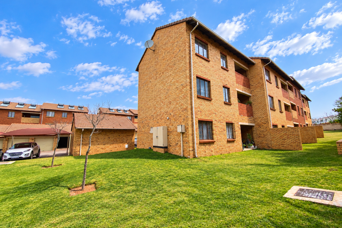 A view of the rear exterior of a face brick townhouse complex featuring a well-maintained lawn and integrated carport structures.