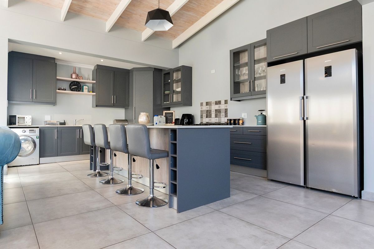 Modern kitchen featuring dark grey cabinetry, a central island with breakfast bar seating, tiled floors, and an exposed beam ceiling.