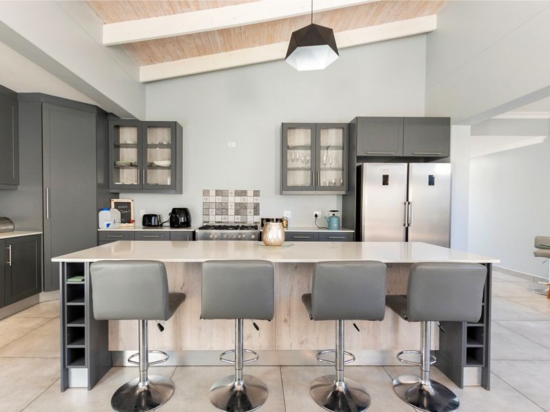 Modern kitchen featuring a large central island with breakfast bar seating, sleek grey cabinetry, quartz countertops, and a high vaulted ceiling with exposed beams.