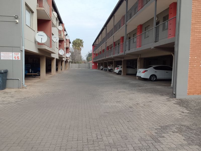 Exterior view of an apartment complex featuring a paved driveway, covered parking bays, and multi-level balconies with brick and painted concrete finishes.