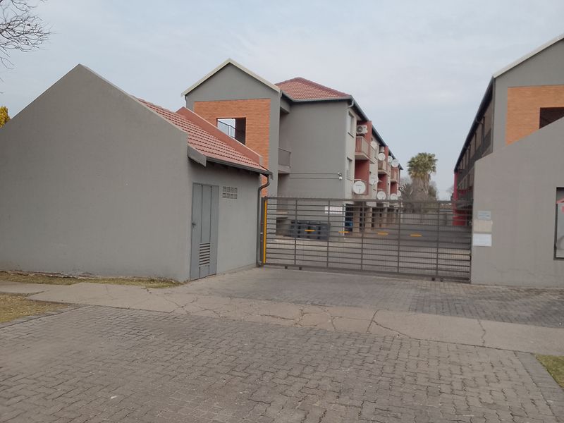 Exterior view of an apartment complex entrance featuring a security gate, paved driveway, and a small utility structure.