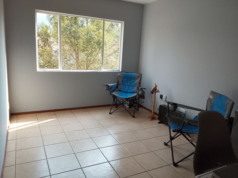 A bright, empty living area featuring neutral tiled flooring and a large window providing natural light and a pleasant outdoor view.