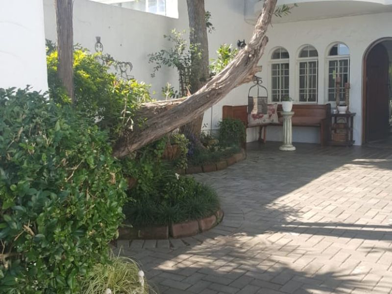 A private paved courtyard featuring mature trees, landscaped garden beds, and an arched entrance leading into the townhouse with an overhead balcony.