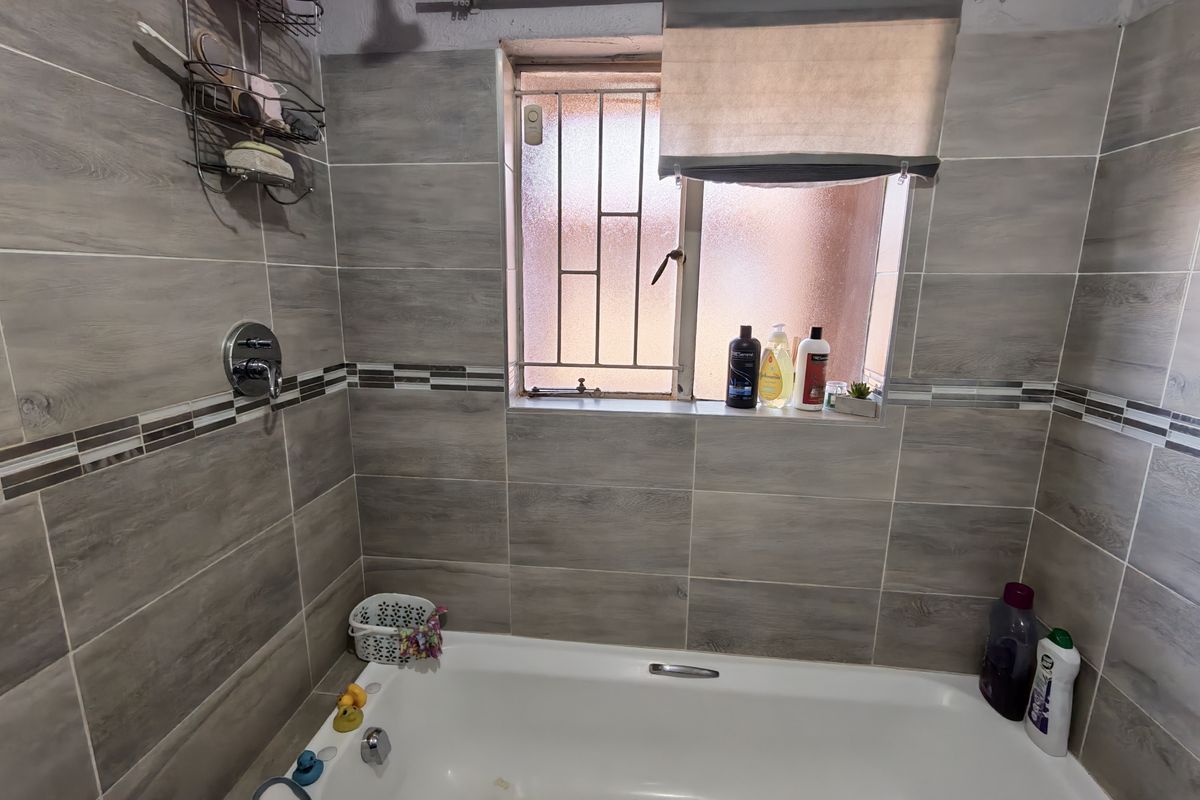 A bathroom featuring a built-in bathtub with shower mixer, tiled walls, and a window with security bars.