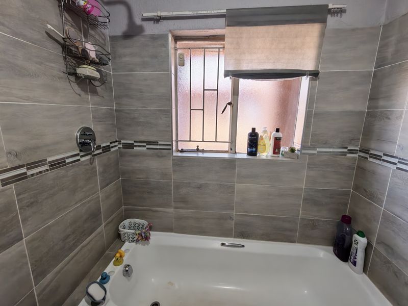 A bathroom featuring a built-in bathtub with shower mixer, tiled walls, and a window with security bars.