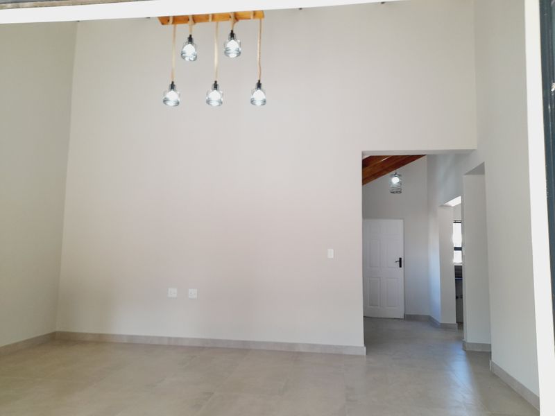 A bright, open-plan living area featuring neutral tiled flooring, high ceilings, and modern pendant lighting, leading into a secondary hallway space.