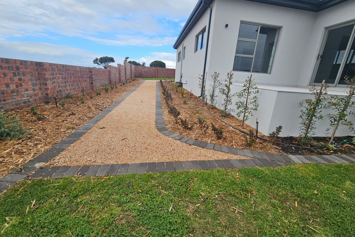 A well-maintained rear garden area featuring a gravel pathway with paver edging, a brick boundary wall, and established young plantings along the house exterior.