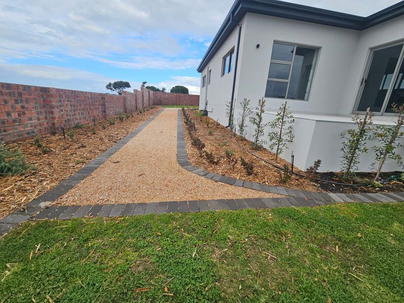 A well-maintained rear garden area featuring a gravel pathway with paver edging, a brick boundary wall, and established young plantings along the house exterior.