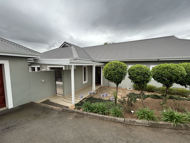 Front exterior view of the townhouse featuring a covered walkway, paved entrance area, and well-maintained landscaped garden with a corrugated metal roof.
