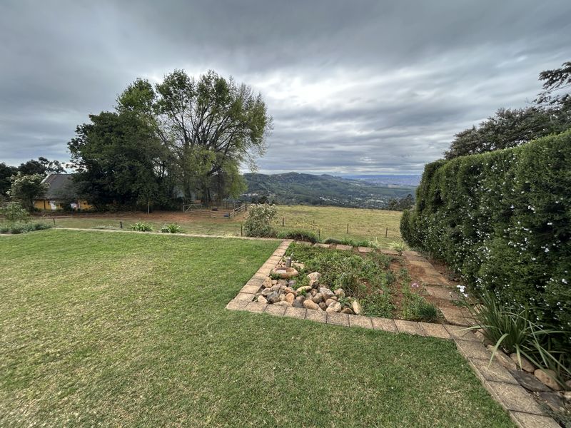 A scenic view overlooking a landscaped garden with stone-edged beds, mature hedging, and expansive rolling hills in the distance.