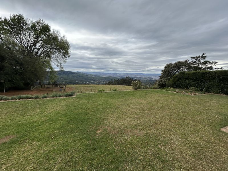 Expansive elevated view overlooking a manicured lawn, surrounding greenery, and distant valley landscape under a cloudy sky.