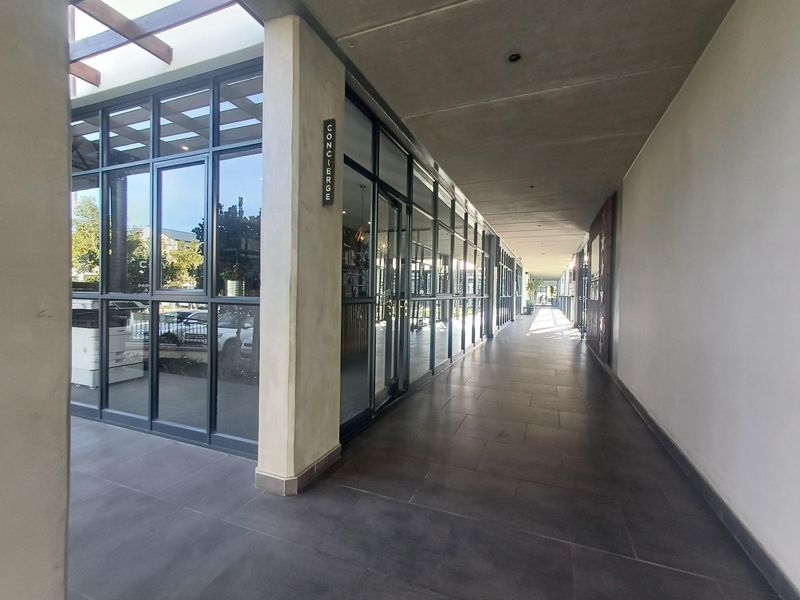 A modern, covered outdoor walkway featuring tiled flooring, concrete ceilings, and a large glass-fronted concierge area.